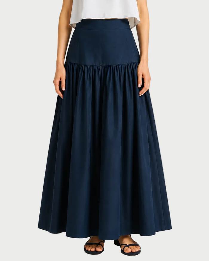 A woman in a white sleeveless top and the Saint Art NY Tossa Poplin Maxi Skirt, featuring a flared, gathered drop-waist silhouette, paired with black sandals, stands against a plain background.