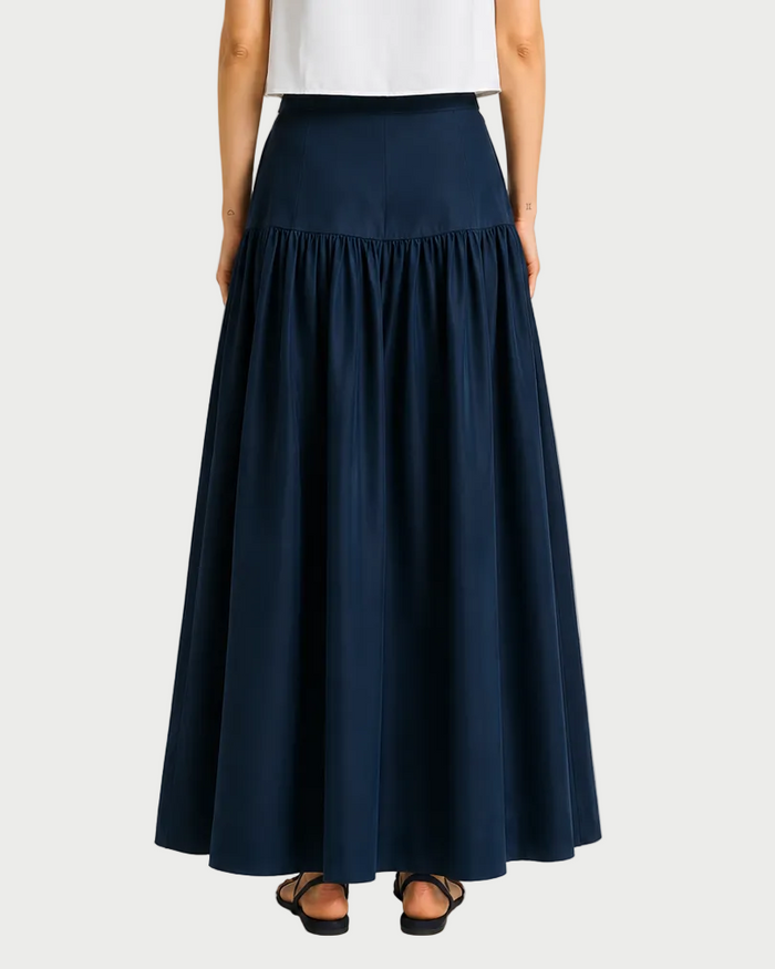 A person shown from the back wears a white top and the Saint Art NY Tossa Poplin Maxi Skirt in dark navy, featuring a gathered fabric drop-waist silhouette to the ankles, paired with black sandals.