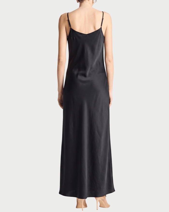 A woman viewed from the back wearing a long black satin slip dress with thin spaghetti straps and nude high heels, set against a solid light gray background.