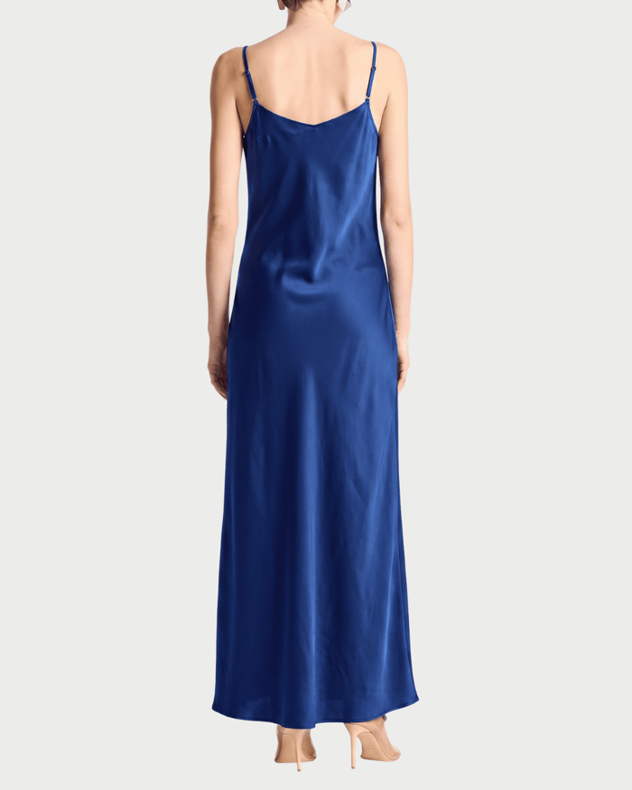 A woman shown from the back wearing a long royal blue satin slip dress with thin spaghetti straps, paired with beige high heels, against a light gray studio background.