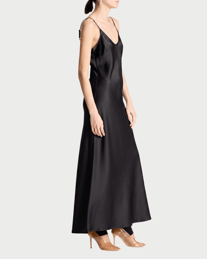 A model wearing a black satin slip dress with thin spaghetti straps and a V-neckline, paired with clear high-heeled pumps, shown from a side profile against a light gray background.