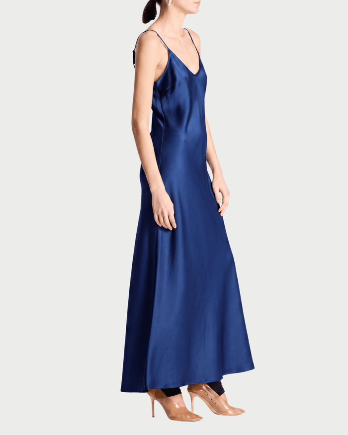 A woman modeling a long navy blue satin slip dress with thin straps, shown in profile against a light gray studio background.