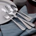 Angled view of Satin Annapolis 45-piece stainless flatware showing satin brushed finish on a blue napkin beside a white plate