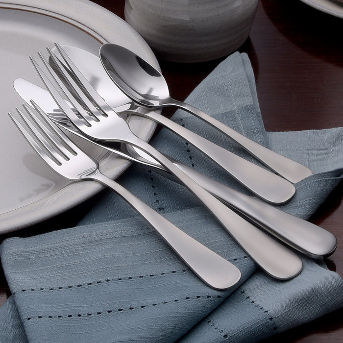 Angled view of Satin Annapolis 45-piece stainless flatware showing satin brushed finish on a blue napkin beside a white plate