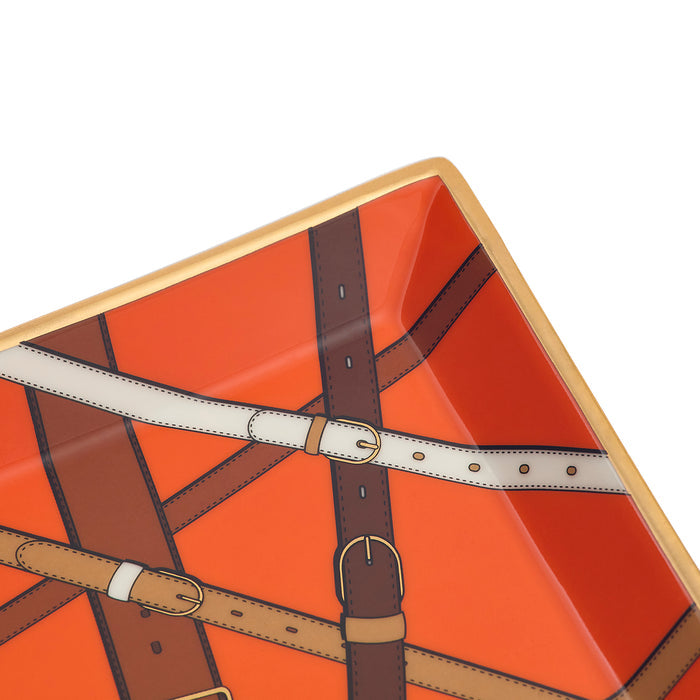 Close-up of a glossy orange tray or plate with an illustrated leather-buckle strap pattern in brown, tan, cream, and gold-toned edging, photographed on a white background.