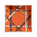 A square ceramic plate featuring a vibrant orange background with a printed pattern of intersecting leather straps in brown, tan, and white, framed by a light beige rim.