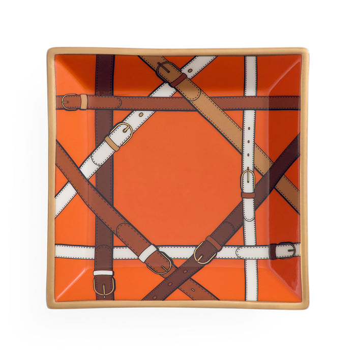 A square ceramic plate featuring a vibrant orange background with a printed pattern of intersecting leather straps in brown, tan, and white, framed by a light beige rim.