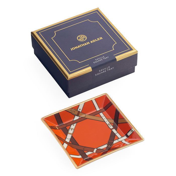 Jonathan Adler Savile Square Tray shown with its navy blue and gold presentation box on a white background. The tray features a vibrant orange background with brown leather-strap patterns and white details.