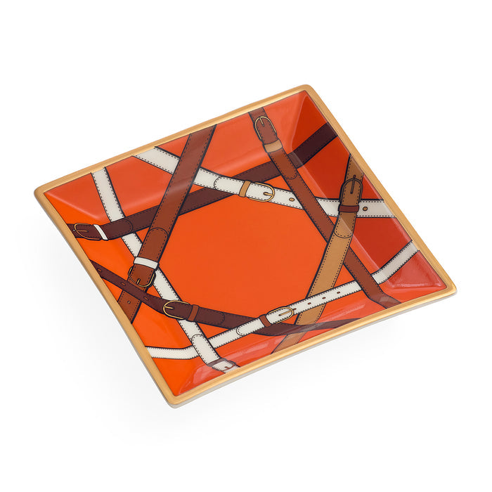 A square ceramic dish featuring a vibrant orange and red background with an artistic print of leather straps and buckles in brown, beige, and white, finished with a light gold rim.