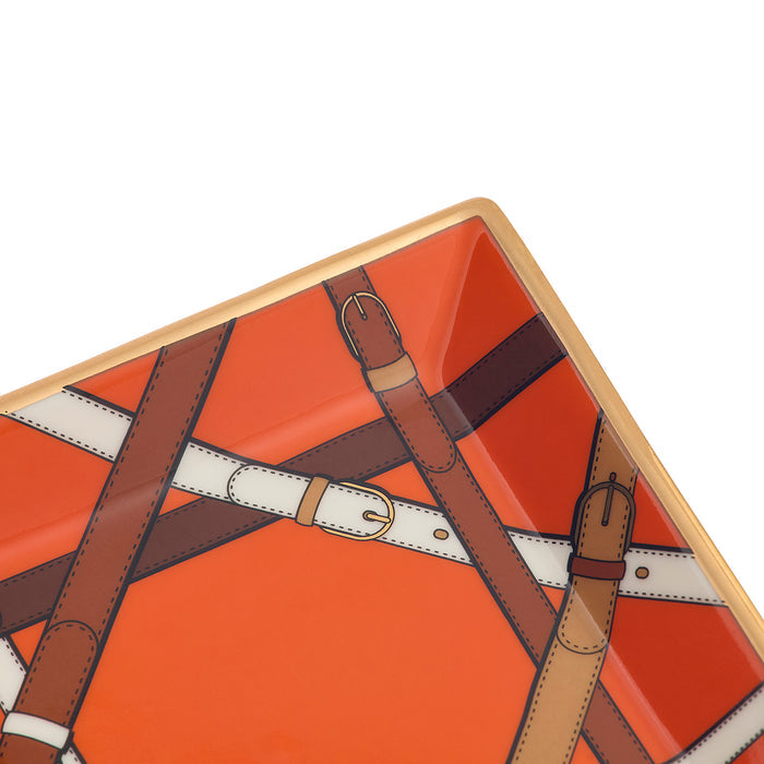 Close-up of a square decorative tray with rounded corners, featuring an orange background and an illustration of brown and white leather straps with buckles, finished with a gold-toned rim.