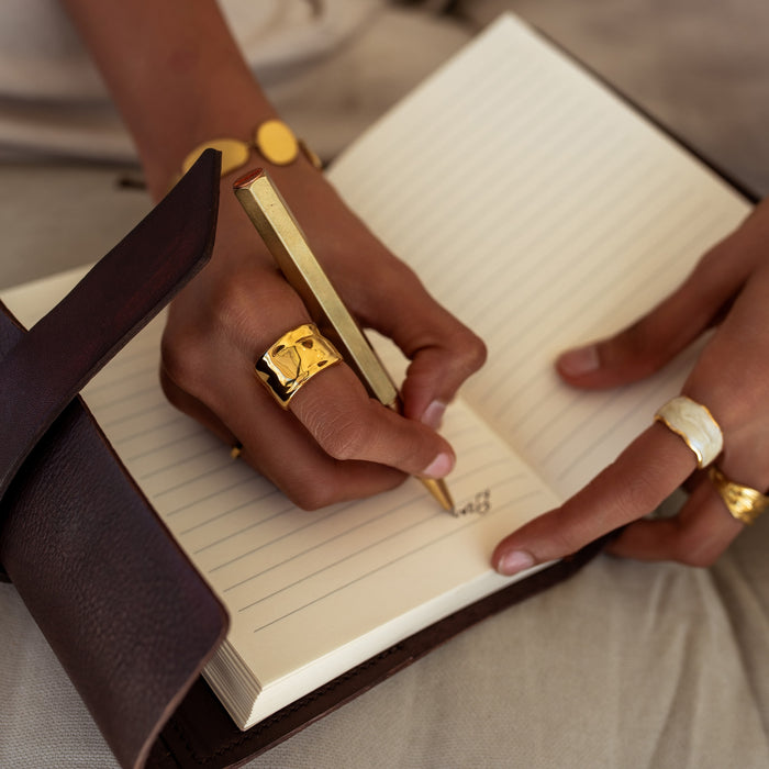 Person writing in a notebook with a gold pen and rings on fingers.