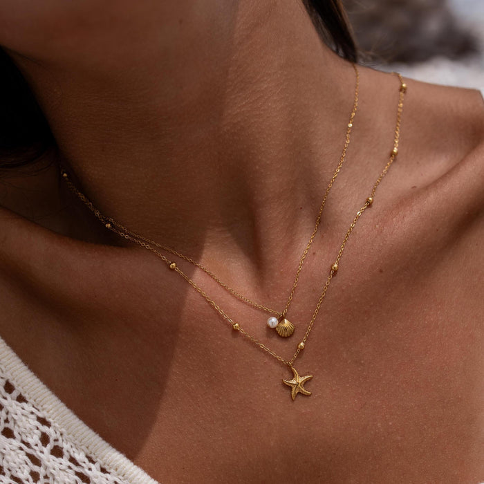 Close-up of a person wearing a dainty gold double layer necklace with starfish, shell, and pearl pendants.