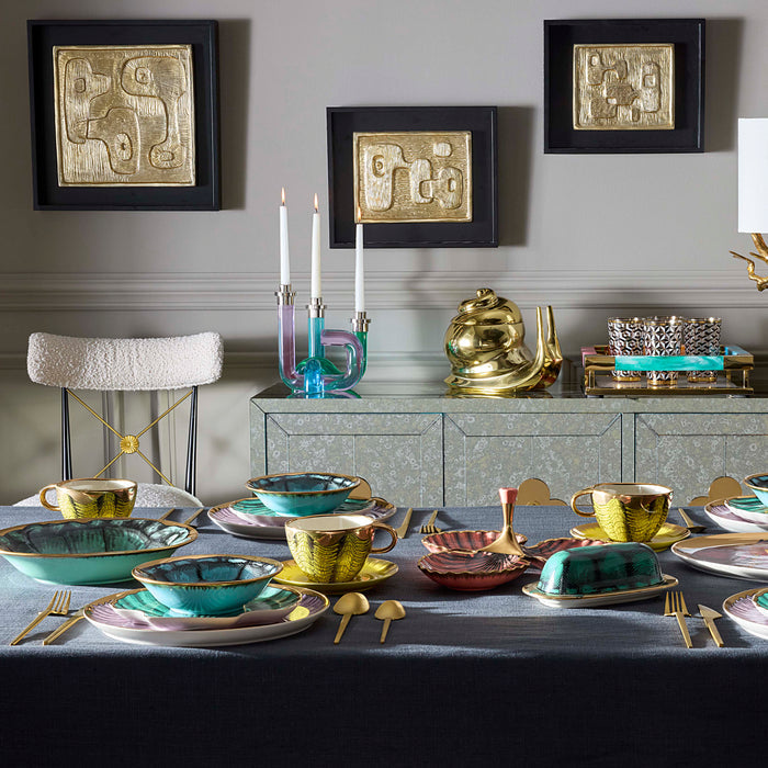 A styled dining table set with gold and turquoise ceramic tableware, including teacups, plates, and butter dishes, alongside matching gold and turquoise candlesticks and a gold teapot on a mirrored sideboard. Three framed abstract gold artworks hang on a gray wall above.