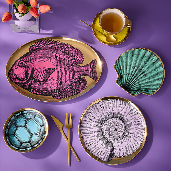 A styled flat-lay of a dinnerware set on a purple background, featuring a large oval platter with a pink fish illustration, a scallop-shaped dish in teal, a bowl with a blue soccer pattern, and two round plates with gold-leaf-edged nautilus and spiral designs. Gold cutlery, a yellow teacup, and a vase of tulips are arranged around them.