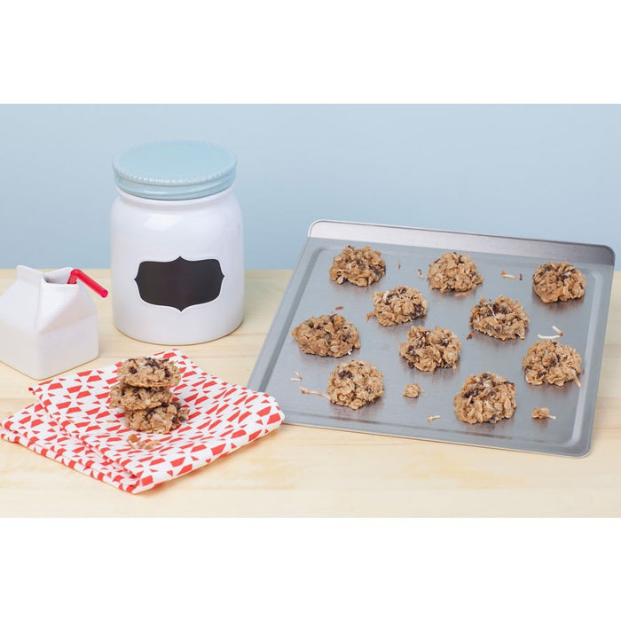 Angled view of medium stainless steel cookie sheet with oatmeal cookies, red-and-white napkin and milk carton on a light wood surface