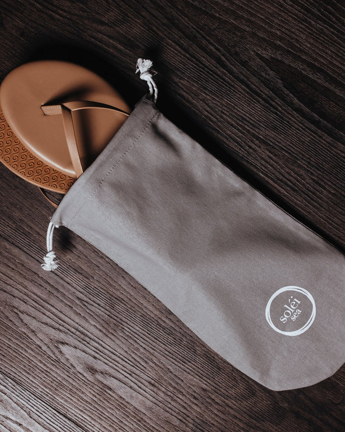 The Solei Sea Indie Flip Flop, a tan vegan leather sandal, is partially tucked in a gray "Solei Sea" logo drawstring bag and displayed on a dark wooden surface.