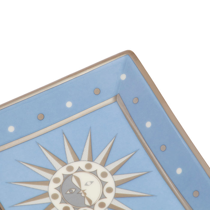 Close-up of a decorative ceramic plate corner featuring a light blue field, white dotted border, and a central sun-and-moon motif in cream, taupe, and gray.