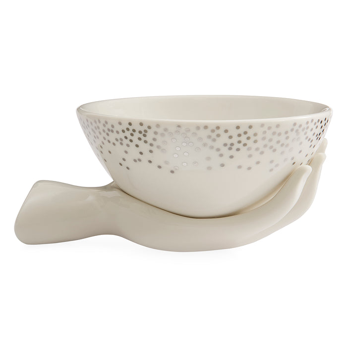 An off-white ceramic bowl with a dotted pattern on the exterior, nestled in a sculptural ceramic hand-shaped base, set against a plain white background.