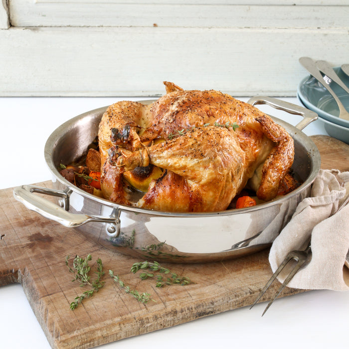 Roasted whole chicken in a stainless-steel roasting pan on a wooden board with thyme, carving fork, and stacked plates.