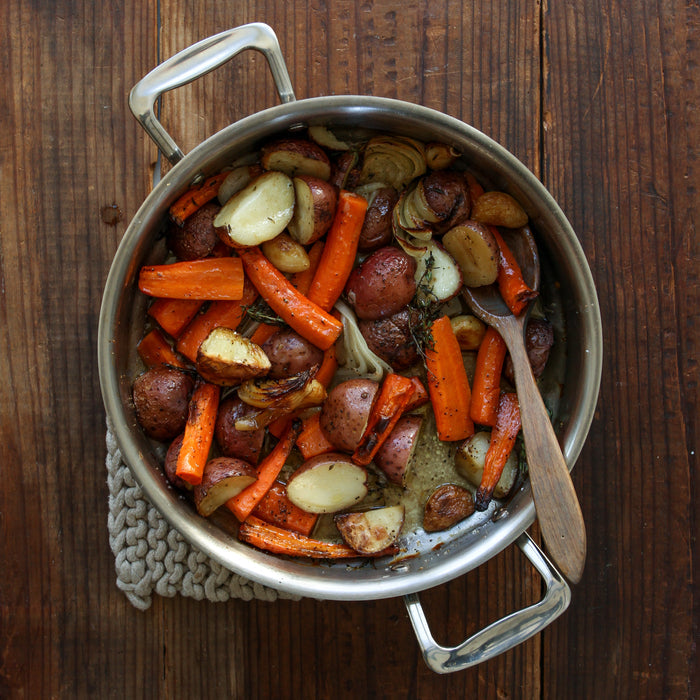 Skillet of roasted baby potatoes, carrots, and onions with a wooden spoon on a wooden table.