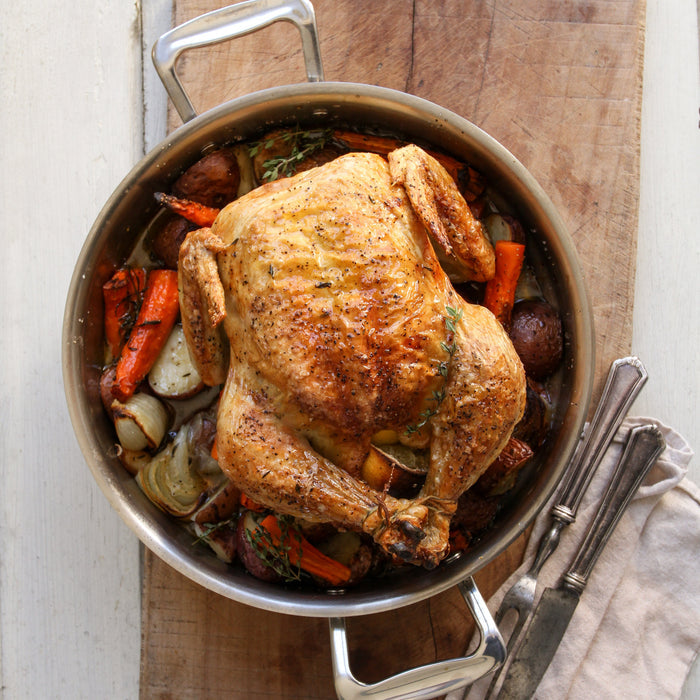 Whole roasted chicken in a stainless-steel pan with carrots, onions and potatoes; serving knife and fork on a cloth.