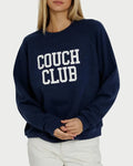 A woman wearing the Sunny Somewhere Couch Club Raglan Crew—a navy blue, oversized raglan sweatshirt with “COUCH CLUB” in white letters on the front—paired with white pants and standing against a plain background.