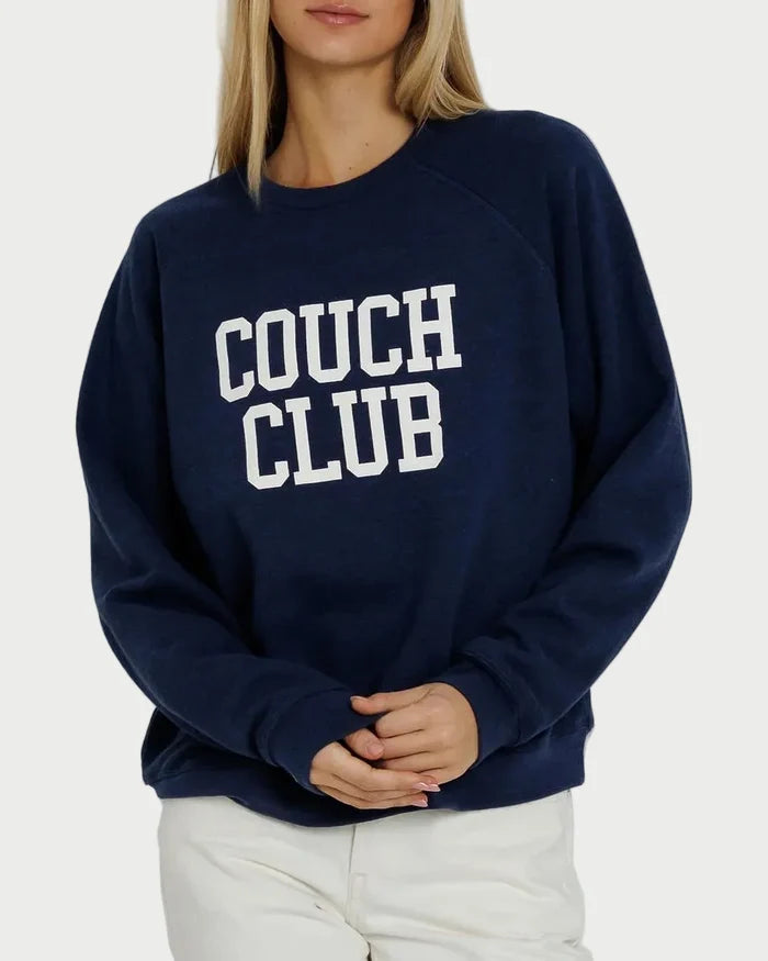 A woman wearing the Sunny Somewhere Couch Club Raglan Crew—a navy blue, oversized raglan sweatshirt with “COUCH CLUB” in white letters on the front—paired with white pants and standing against a plain background.