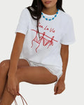 A woman wears the Sunny Somewhere Sea La Vie Tee by Sunny Somewhere—a relaxed-fit white coastal graphic top—paired with lace-trimmed shorts and a blue beaded necklace, sitting comfortably with her hand on her knee.