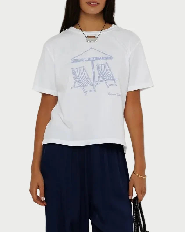 A woman modeling a white short-sleeve graphic T-shirt with a light blue line drawing, paired with dark navy wide-leg pants, black sandals, and holding a black woven handbag against a plain light gray studio background.