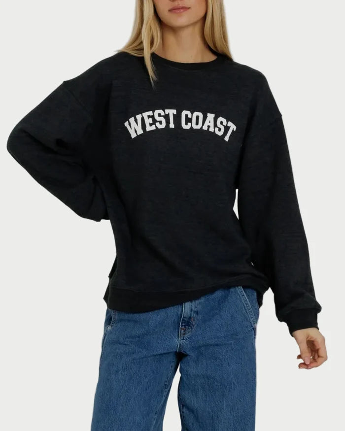A person is wearing the Sunny Somewhere West Coast Oversized Crew by Sunny Somewhere—a black oversized sweatshirt with “WEST COAST” in white letters on the front—paired with blue jeans, standing against a plain background.