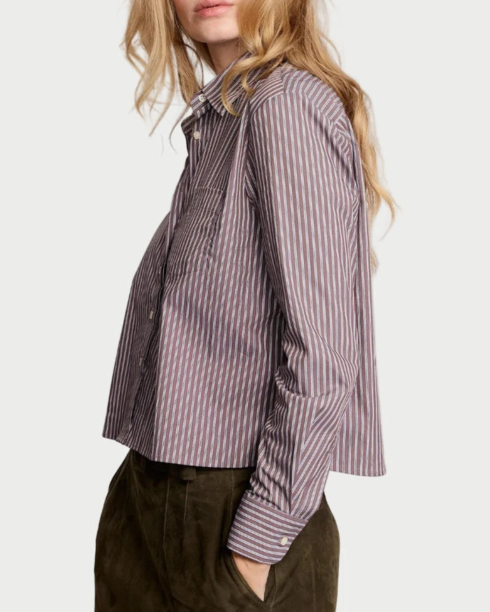 A person with long, wavy blonde hair wears The Shirt Long Sleeve Stripe Boxy Crop by The Shirt in purple and white vertical stripes with No Gape button technology and dark green pants, standing sideways against a plain light background.