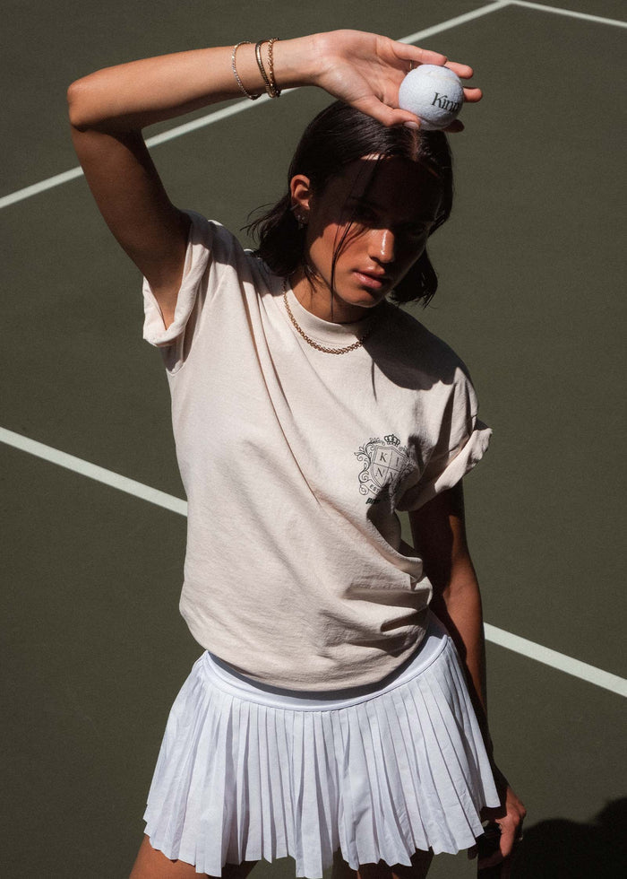 woman on a tennis court wearing a Kinn x Prince The Standard Tee and a white tennis skirt and holding a white tennis ball