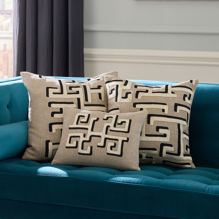 Three beige throw pillows with a black Greek key pattern arranged on a teal tufted sofa in a living room setting.