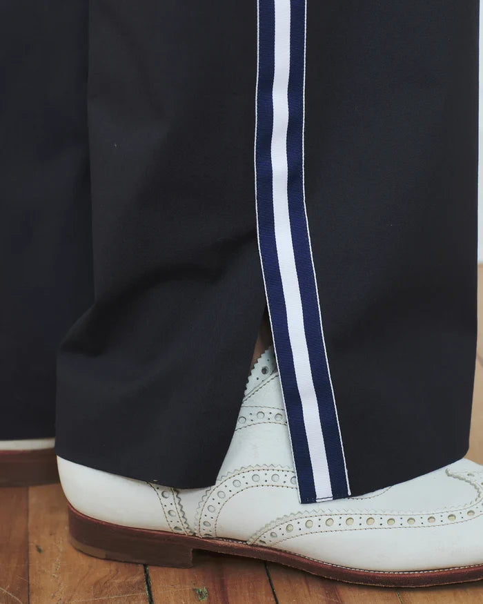 Black pants with white and blue stripes worn with a white shoe on a wooden floor.