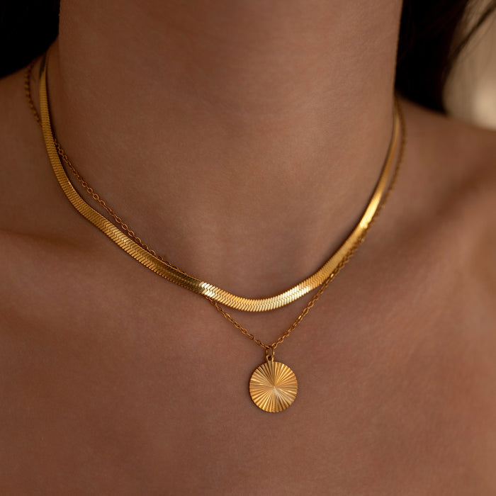 Gold necklace with a circular pendant on a person's neck paired with a sleek gold herringbone necklace against a neutral background