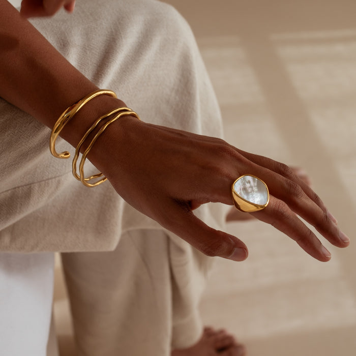 Hand wearing a gold ring with a large mother-of-pearl-like stone set on a person's finger and gold cuffs and bangles on a neutral background