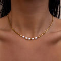 Dainty gold necklace with small square beads and freshwater pearls on a person's neck against a neutral background