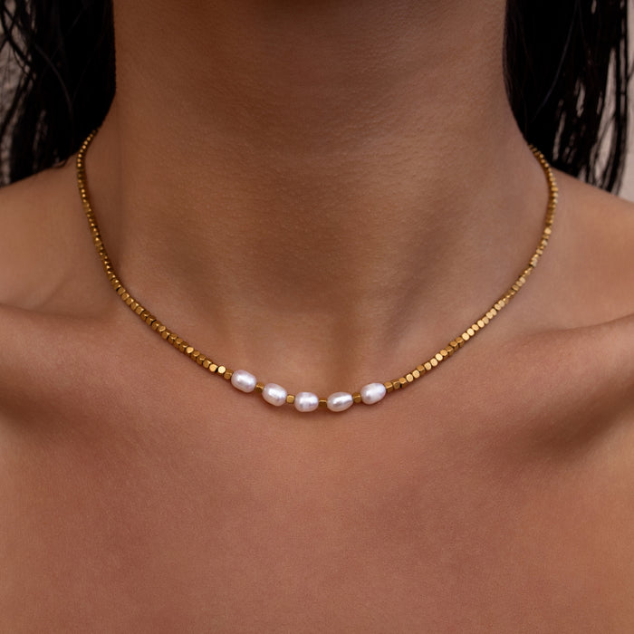 Dainty gold necklace with small square beads and freshwater pearls on a person's neck against a neutral background