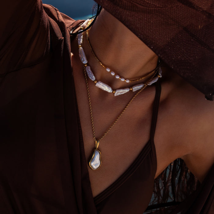 Close up of a woman by the pool wearing a brown bikini and swim dress with 3 gold and pearl necklaces on including a large pearl pendant, long freshwater pearl beads and a dainty gold and pearl bead choker