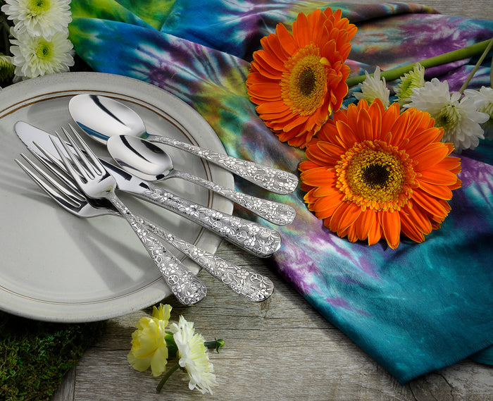 Angled view of Woodstock 45-piece flatware with ornate embossed peace and flower motifs on a plate and tie-dye cloth