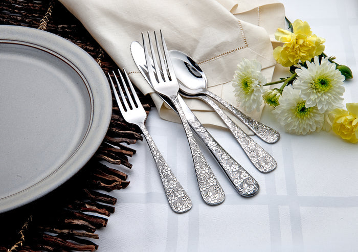 Gray plate with ornate-handled forks and spoons on a napkin beside yellow and white mums on a white tablecloth.