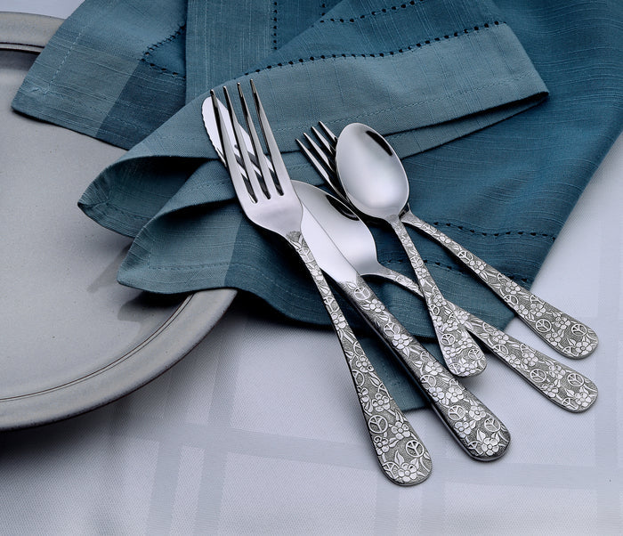 Ornate floral-engraved stainless flatware (forks, spoons) arranged on a blue cloth napkin beside a gray plate.