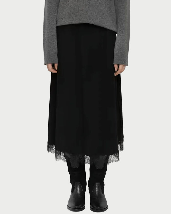 A model stands against a light gray studio background wearing a gray knit sweater, a long black skirt with a sheer tulle hem, black leggings, and black ankle boots.
