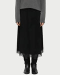 A model stands against a light gray studio background wearing a gray knit sweater, a long black skirt with a sheer tulle hem, black leggings, and black ankle boots.