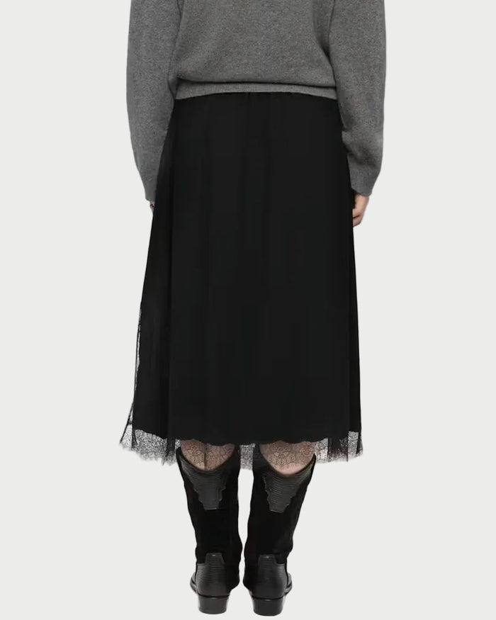 A woman viewed from behind wearing a gray knit sweater, a black midi skirt with sheer tulle hem, sheer black tights, and black ankle boots, against a light gray studio background.